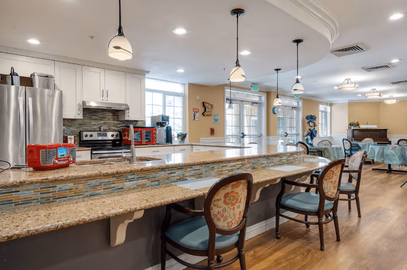 A bright and spacious kitchen and dining area with a long granite countertop island featuring a mosaic tile backsplash. The kitchen has white cabinets, stainless steel appliances including a refrigerator and stove, and a red retro-style radio on the counter. There are several pendant lights hanging from the ceiling and floral upholstered chairs lined up along the counter. In the background, there are round dining tables with chairs covered in light blue tablecloths, large windows, and double glass doors leading outside.