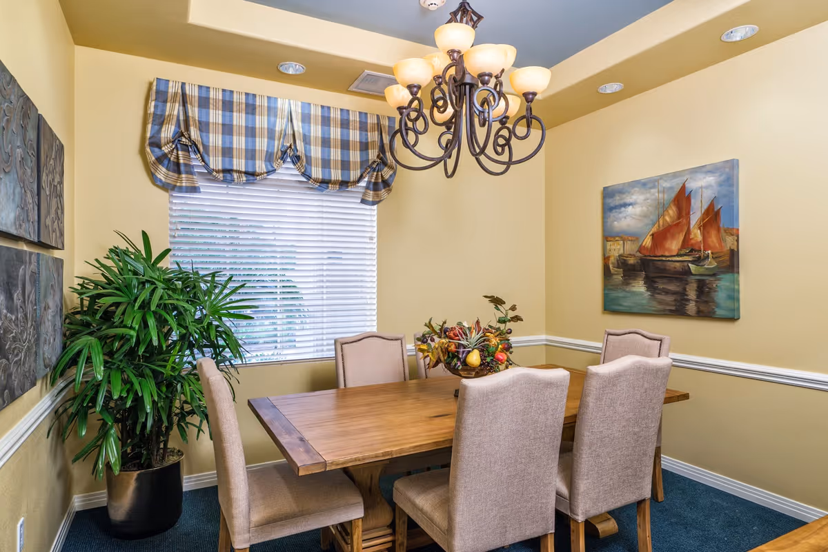 A dining room with a wooden table surrounded by six upholstered chairs. A decorative centerpiece with artificial fruits is on the table. The room has yellow walls with white trim, a window with white blinds and a blue and beige plaid valance. There is a large green potted plant in the corner and two paintings on the walls, one depicting a sailboat scene. A chandelier with multiple lights hangs above the table.