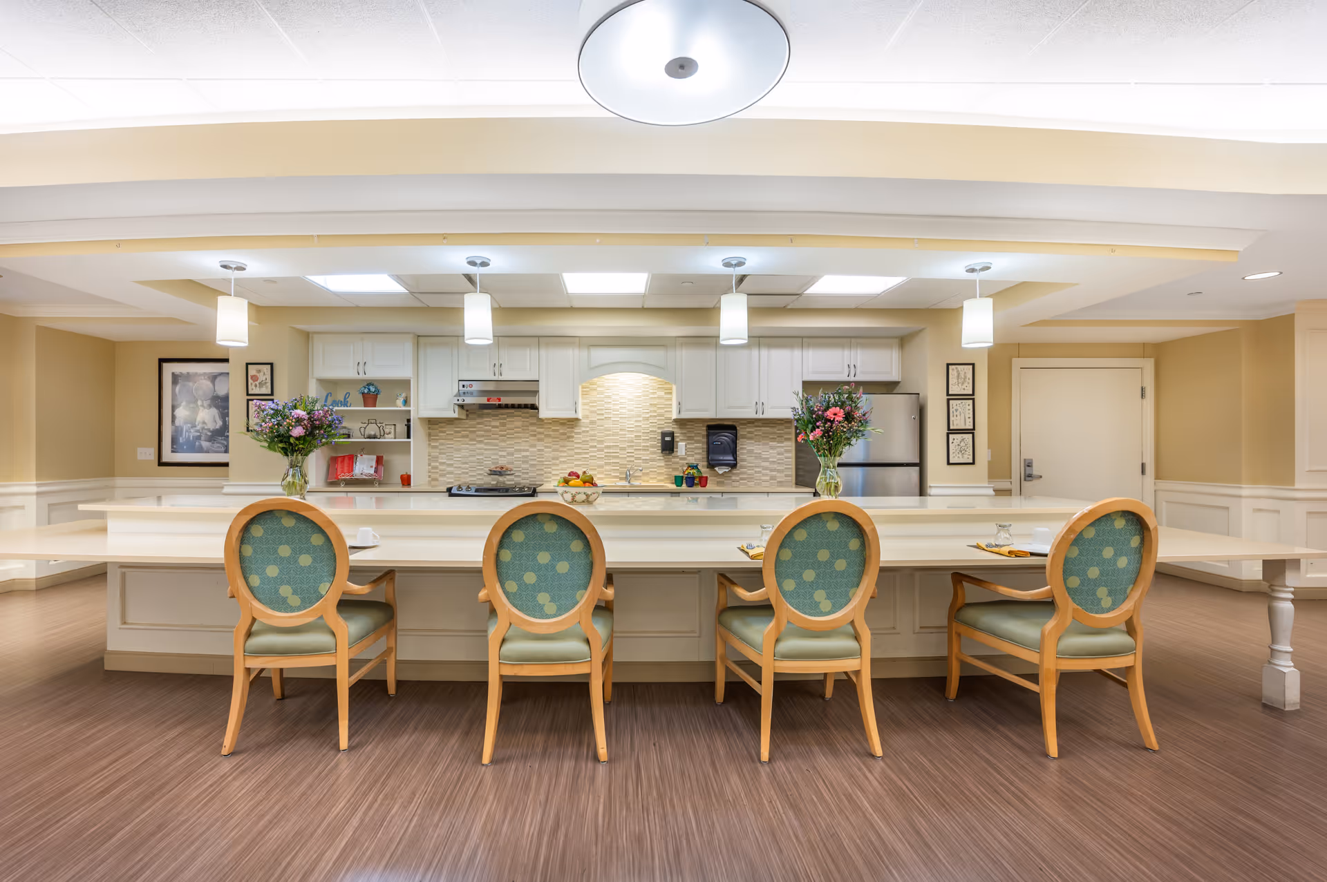 Bright communal kitchen and dining counter with four upholstered chairs, pendant lights, and vases of flowers.