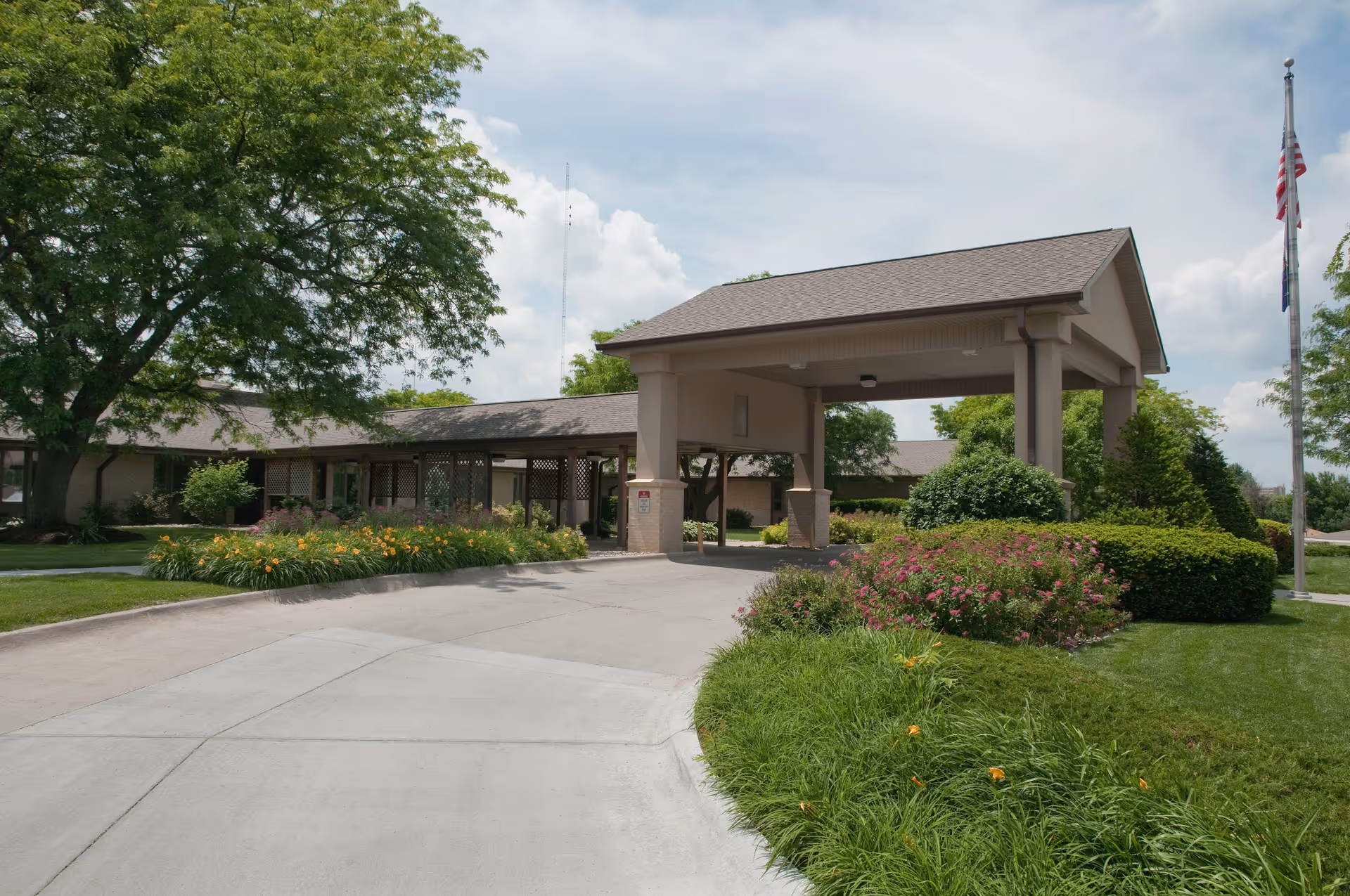 Entrance driveway to a single-story building with a covered drop-off area, surrounded by well-maintained landscaping including green grass, bushes, flowering plants, and a large tree. An American flag is visible on a flagpole to the right.