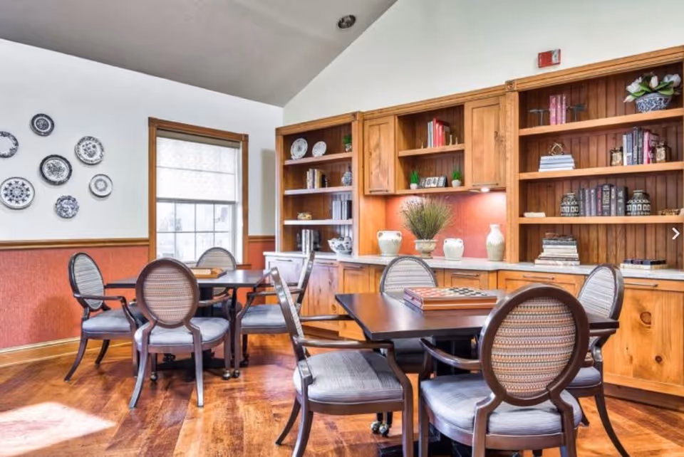 A cozy interior room with wooden flooring and wooden built-in shelves filled with books and decorative items. There are two dark wooden tables, each surrounded by four cushioned chairs with patterned upholstery. A window with a blind lets in natural light, and decorative plates are hung on the white wall above a red wainscoting.