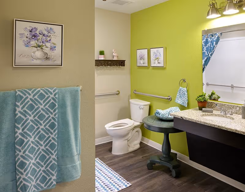A bathroom with a white toilet, a green wall with two framed floral pictures, a granite countertop with a sink, a round green side table holding folded towels, a blue patterned shower curtain, and blue towels hanging on a rack. The floor is dark wood, and there is a small shelf with decorative items above the toilet.