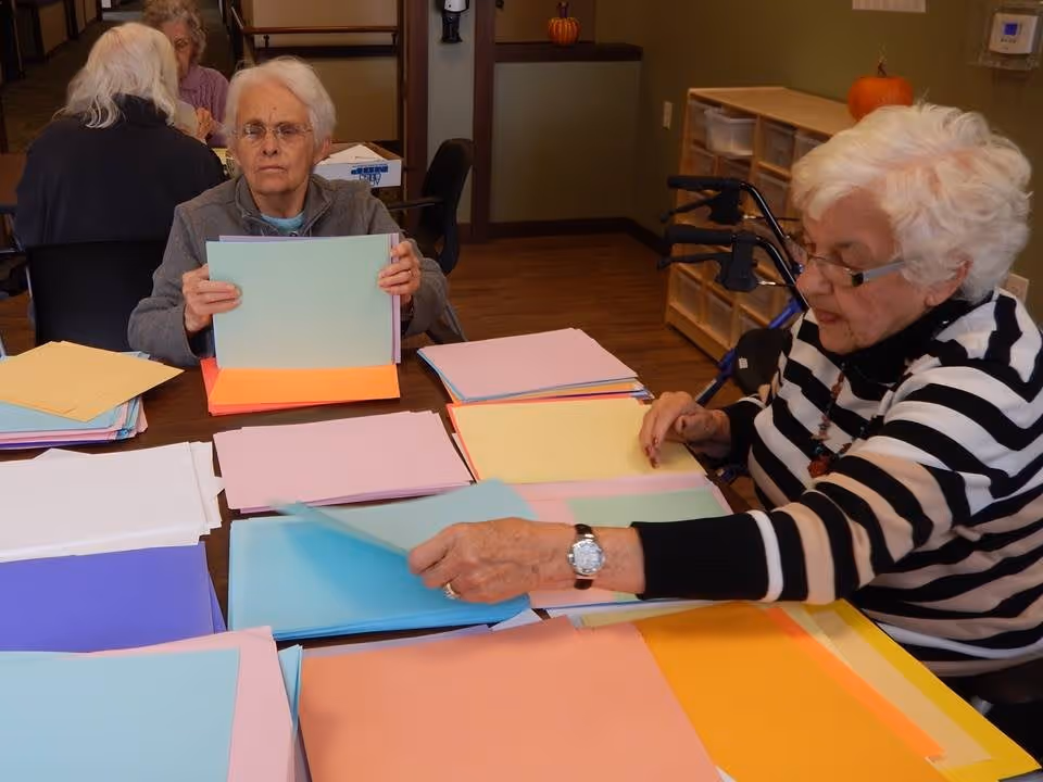 Two elderly women sitting at a table sorting and organizing stacks of colorful paper in a room with wooden flooring and shelves. One woman is wearing a gray jacket and glasses, holding a stack of light blue paper, while the other woman, wearing a black and white striped sweater and glasses, is reaching for a sheet of blue paper. In the background, there are two more elderly women sitting at another table.