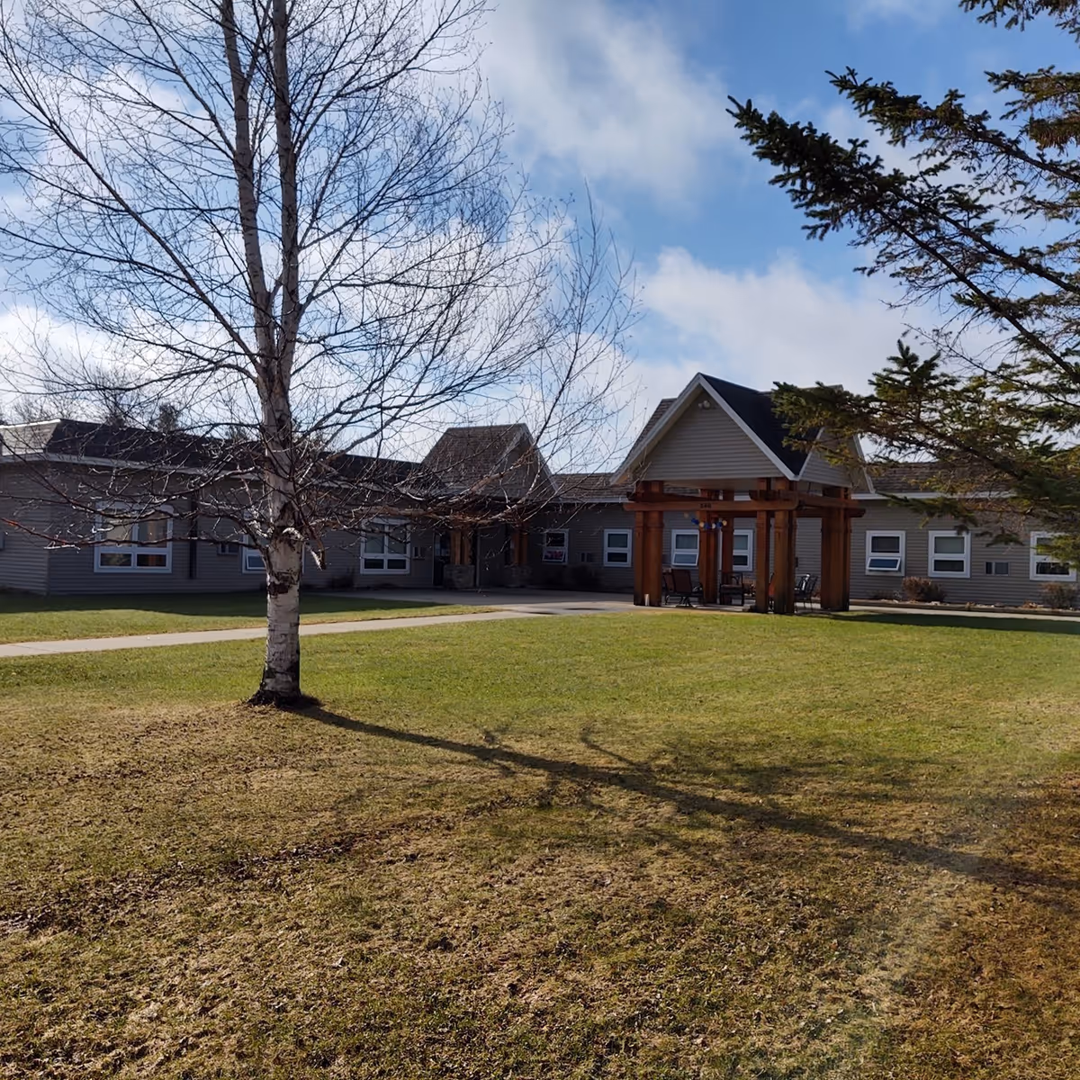 Exterior view of a single-story building with a peaked roof and a wooden pergola structure at the entrance. The building is surrounded by a grassy lawn with a few trees, one of which is leafless, and a partly cloudy sky overhead.