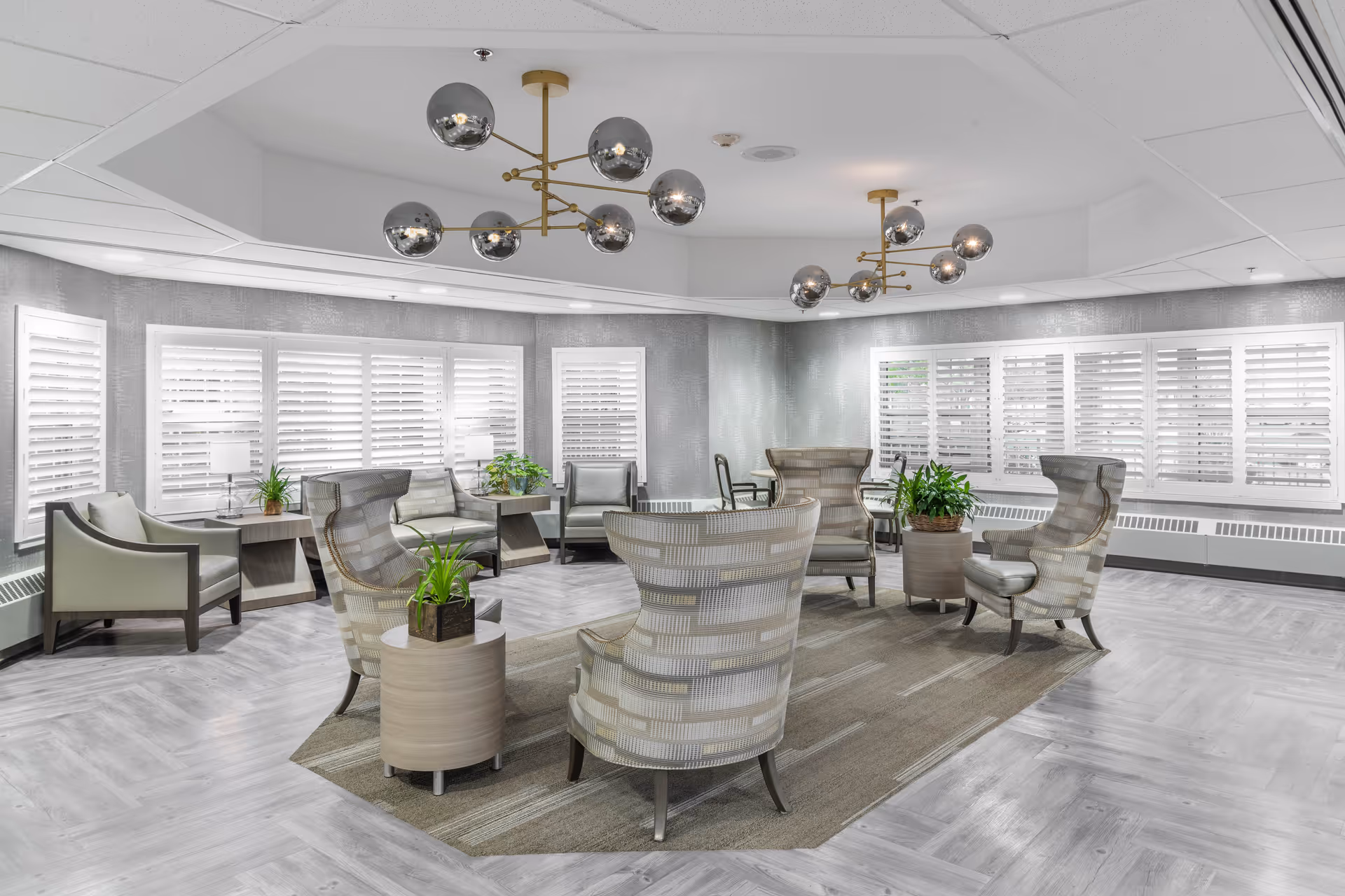 A bright and spacious common area with modern furniture including several armchairs arranged around small tables with potted plants. The room features large windows with white plantation shutters, light gray walls, and two contemporary chandeliers with multiple spherical lights hanging from the ceiling. The floor has a light wood pattern with a large area rug in the center.