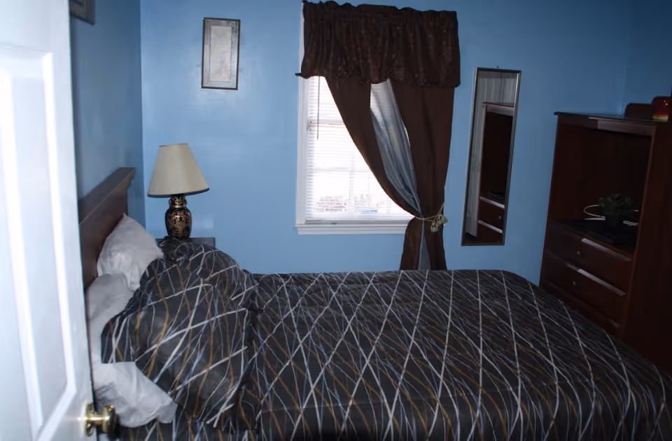 A small bedroom with light blue walls, a bed with a dark patterned comforter, a bedside table with a decorative lamp, a window with brown curtains, a wall-mounted mirror, and a wooden dresser with a small plant on top.