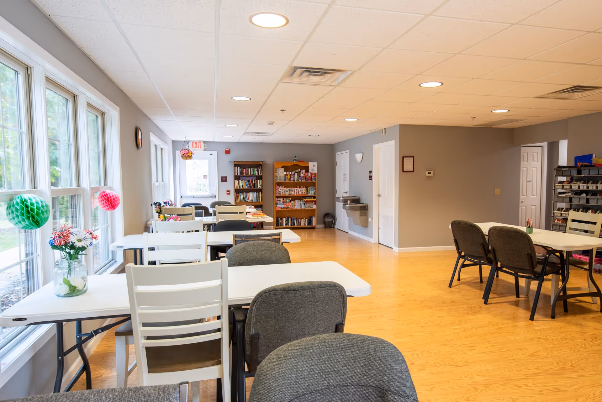 A bright and spacious common area with several white tables and a mix of white and gray chairs arranged around them. Large windows on the left side let in natural light, and colorful paper decorations hang near the windows. In the background, there are bookshelves filled with books and games, a water fountain, and a door with an exit sign above it. The floor is light wood, and the walls are painted gray.