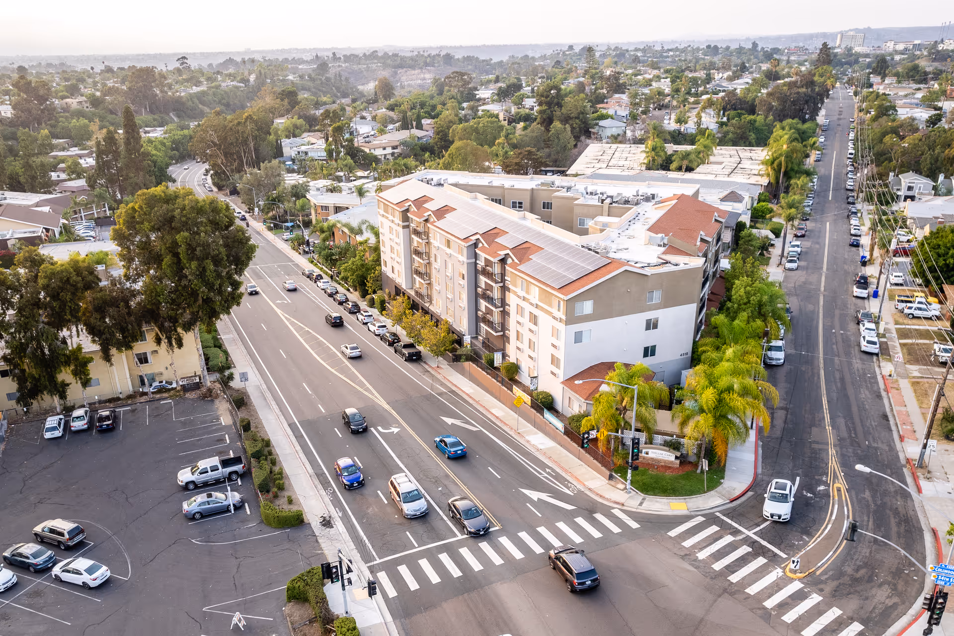 Aerial view of a multi-story residential building at a busy intersection with cars, palm trees, and surrounding neighborhood.