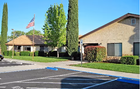 Front exterior of a single-story senior living facility with an American flag, landscaped lawn, and adjacent parking spaces.