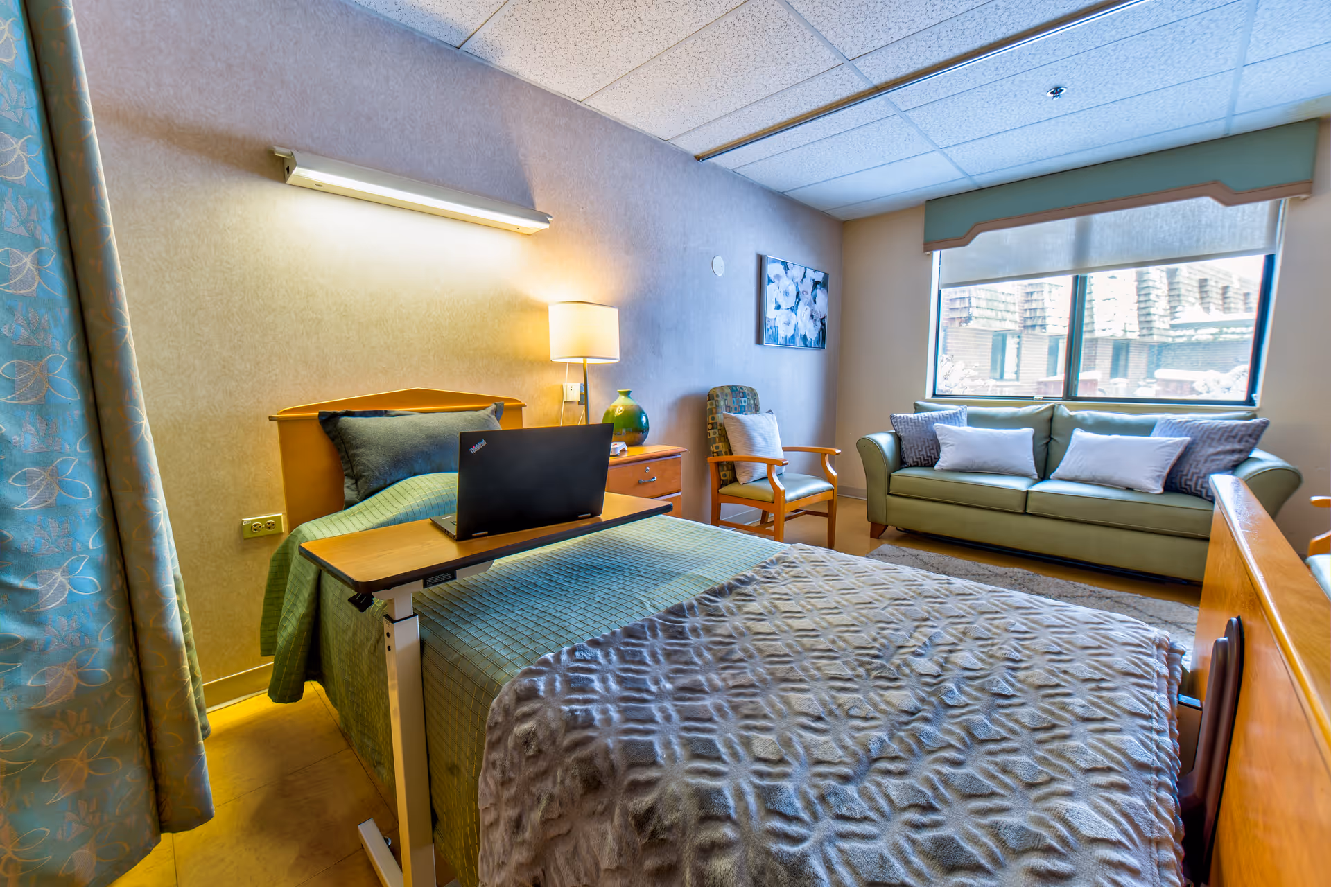 A cozy senior living bedroom with a single bed covered in a green blanket and a gray textured throw. A small overbed table holds a laptop. Next to the bed is a wooden nightstand with a lamp and a green vase. A wooden armchair with a cushion is positioned near the nightstand. Across from the bed is a green sofa with several pillows, placed in front of a large window with a view of an exterior building. The room has soft lighting and a calming atmosphere.