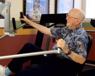 An elderly man wearing a patterned shirt and glasses is exercising on a seated rowing machine in a fitness room with colorful walls and a framed artwork in the background.