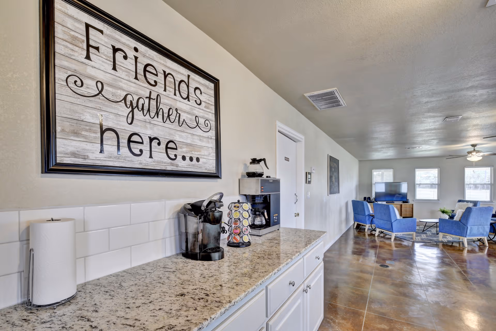 Community lounge with a granite countertop coffee station under a 'Friends gather here...' sign and a seating area with blue chairs and a TV.