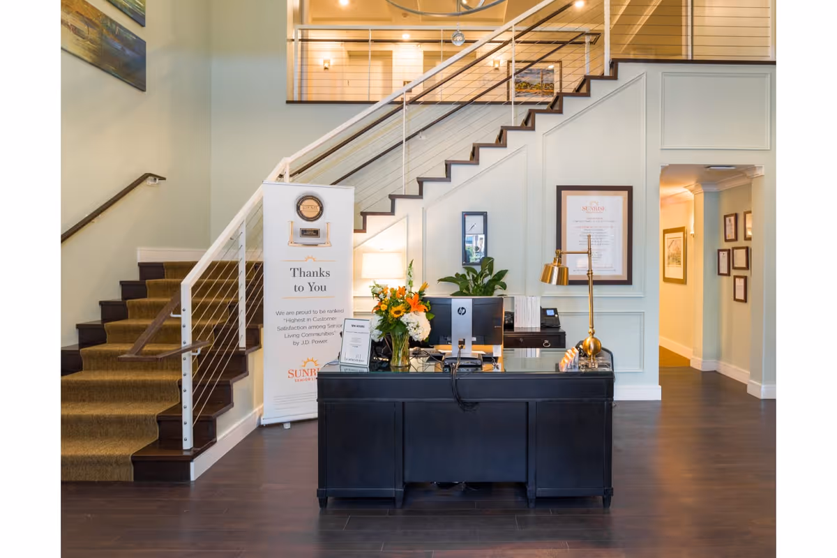 Reception area inside Sunrise of Chevy Chase featuring a black desk with a computer, a gold desk lamp, and a vase of flowers. Behind the desk is a staircase with a modern railing, a banner with a thank you message, framed certificates on the wall, and a hallway leading to other rooms.