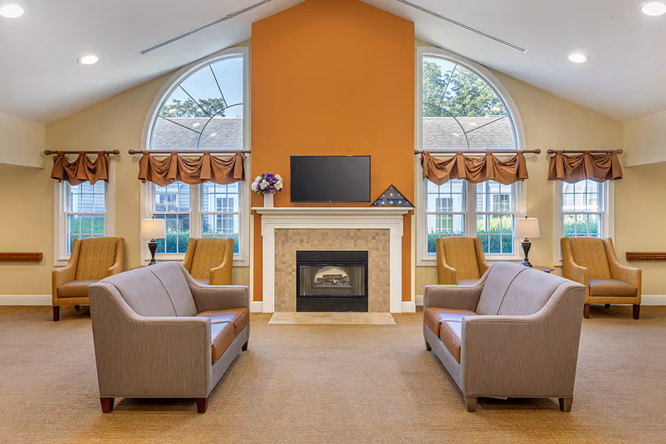 Spacious communal lounge with two sofas facing a fireplace and wall-mounted TV, flanked by armchairs beneath arched windows.