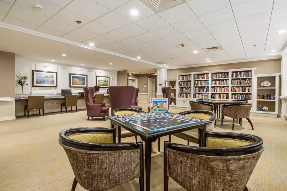 A spacious and well-lit common area in a senior living facility featuring a puzzle table with four chairs in the foreground, a bookshelf filled with books along the back wall, two purple armchairs, and a row of desks with chairs and framed paintings on the left side.