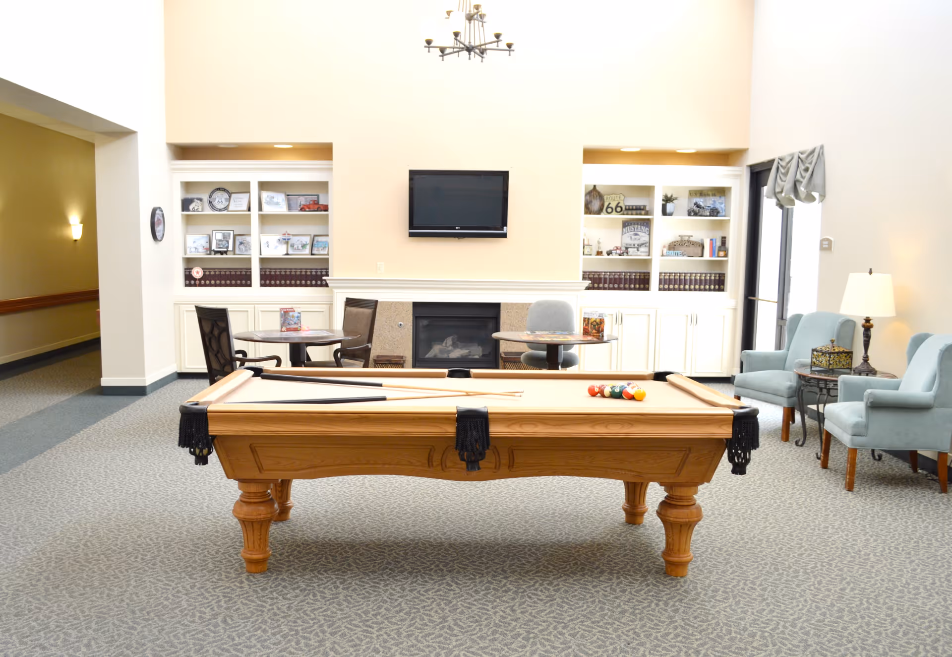 A bright and spacious common area in an assisted living facility featuring a pool table in the center with pool cues and balls arranged on it. Behind the pool table is a fireplace with a flat-screen TV mounted above it, flanked by built-in white shelves filled with books, framed photos, and decorative items. To the right, there are two light blue armchairs with a small side table and lamp between them. To the left, there are two round tables with chairs. The room has carpeted flooring and soft lighting, creating a cozy and inviting atmosphere.