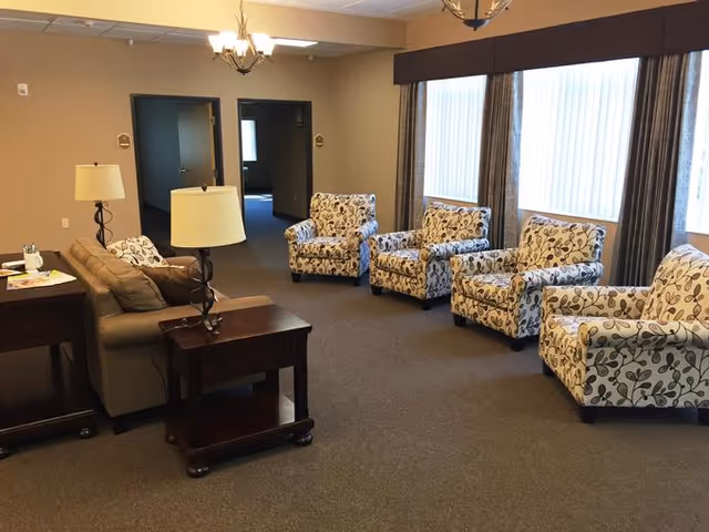 A spacious living room area in an assisted living facility with a beige sofa, four patterned armchairs arranged in a row, two wooden side tables with lamps, and large windows with vertical blinds and curtains allowing natural light to enter.