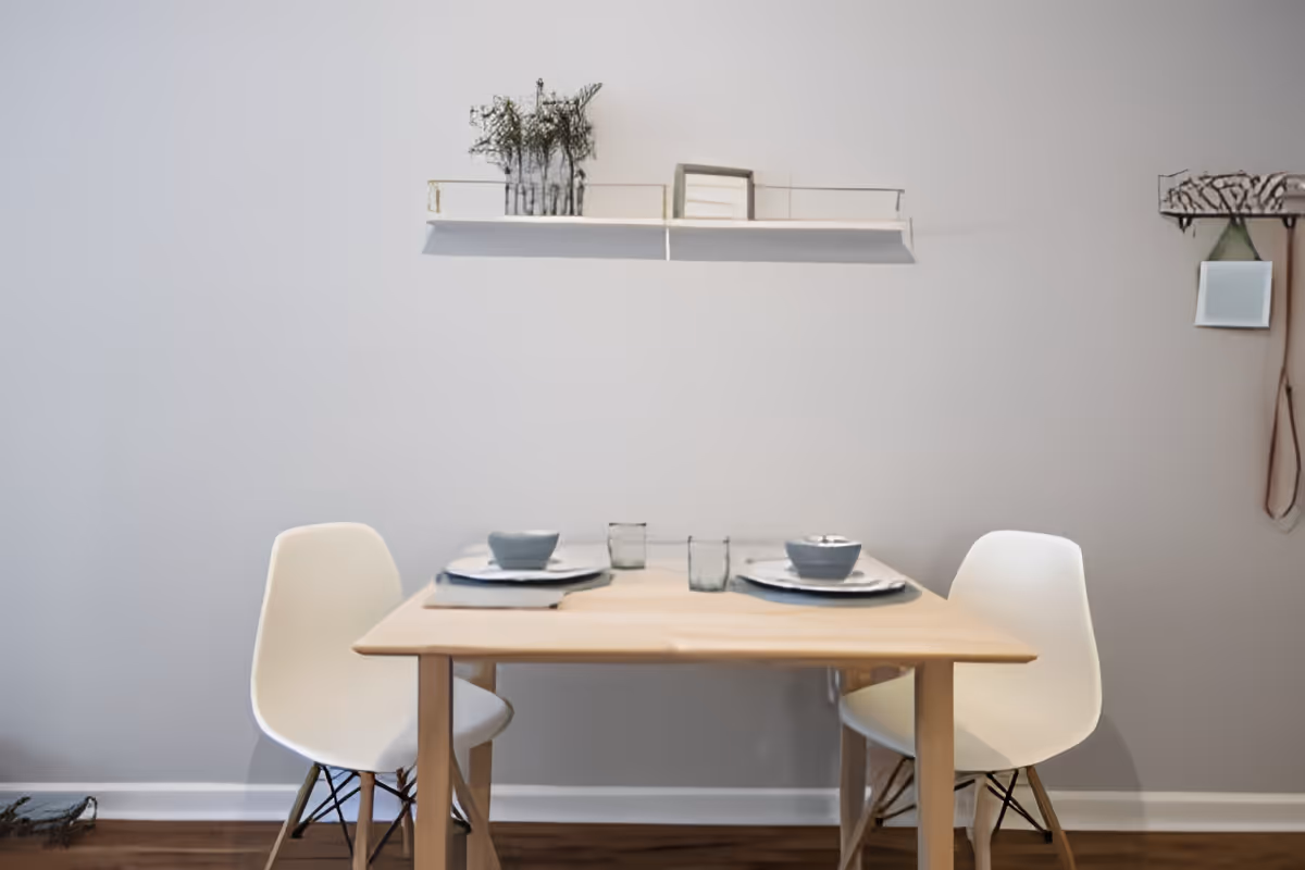 A small dining area with a light wooden table set for two, featuring two white modern chairs. On the table are two bowls, two glasses, and placemats. Above the table is a white wall-mounted shelf with decorative items including small plants and a framed picture. To the right on the wall is a small hanging mirror and a decorative hook with a strap.