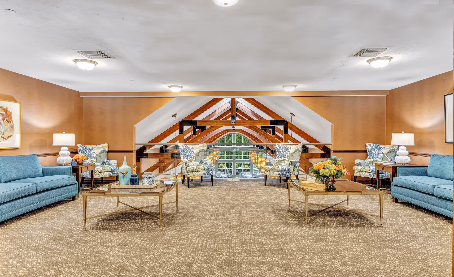 A spacious and well-lit living room area with two teal sofas and four floral-patterned armchairs arranged around two glass coffee tables. The room features warm brown walls, carpeted flooring, and decorative lamps on side tables. Large wooden beams and a glass railing overlook a bright area with large windows and hanging pendant lights.