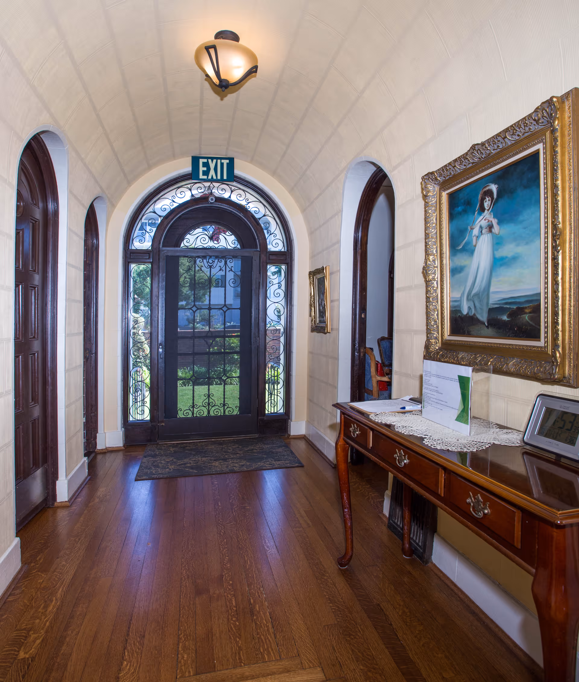 A hallway with a wooden floor and arched ceiling leading to a decorative black wrought iron door with glass panels. Above the door is a green exit sign. On the right side of the hallway, there is a wooden console table with a lace runner, a framed painting of a woman in a white dress, and a digital clock. The walls are light-colored, and there are arched doorways on both sides of the hallway.