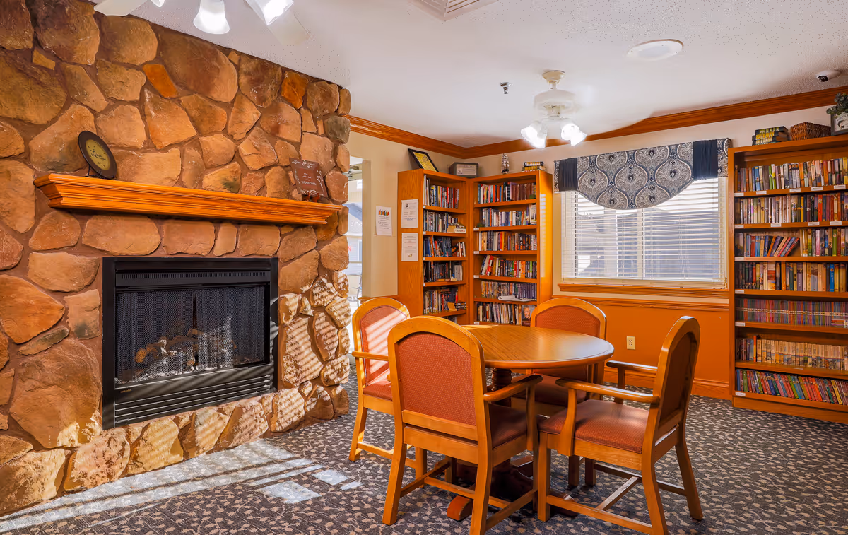 Cozy common room with a stone fireplace, round wooden table and chairs, and bookshelves by a window.