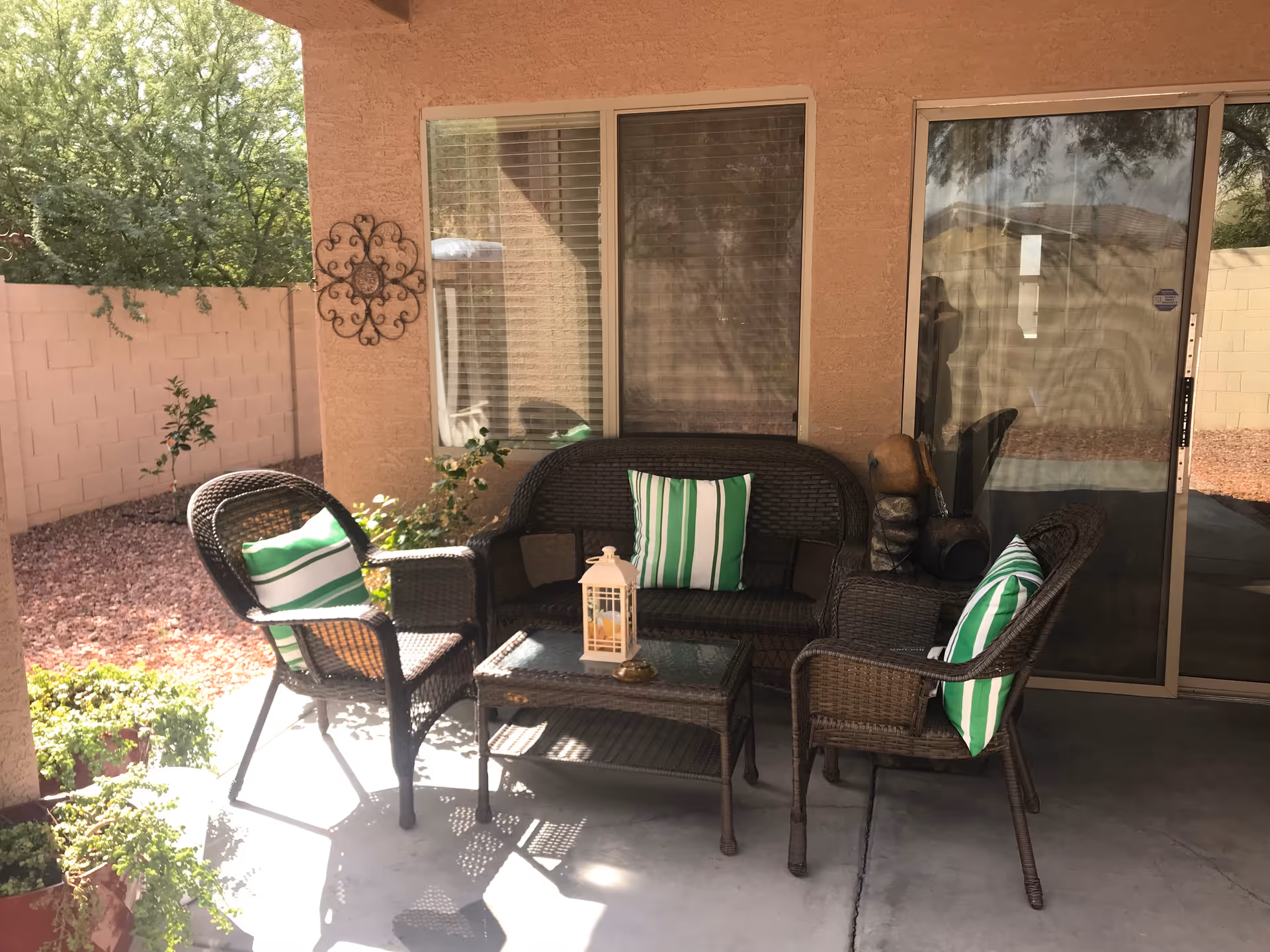 Covered backyard patio with wicker sofa and chairs around a glass-top table, featuring green-striped pillows and potted plants.