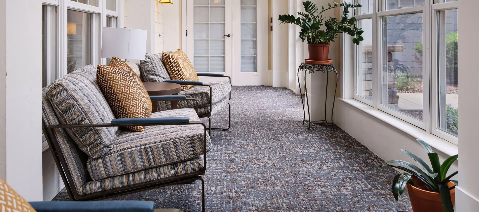 A bright sunroom-style sitting area with striped armchairs and patterned pillows arranged along large windows, small side tables, potted plants, and a carpeted floor.