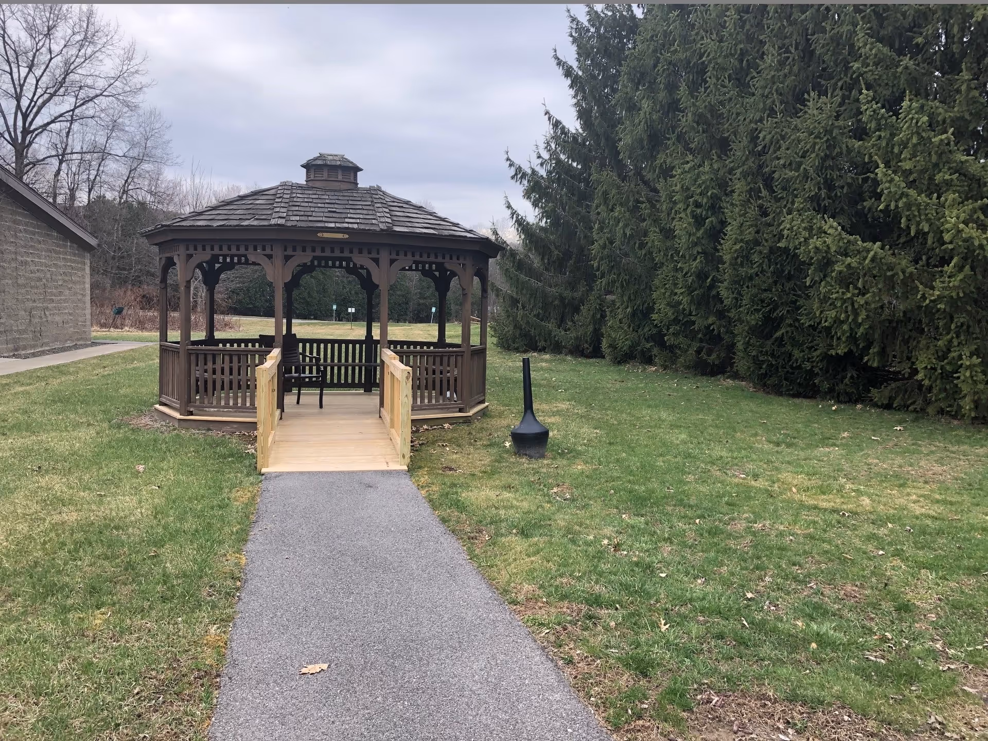 A wooden gazebo at the end of a paved path on a grassy lawn with tall evergreen trees in the background.