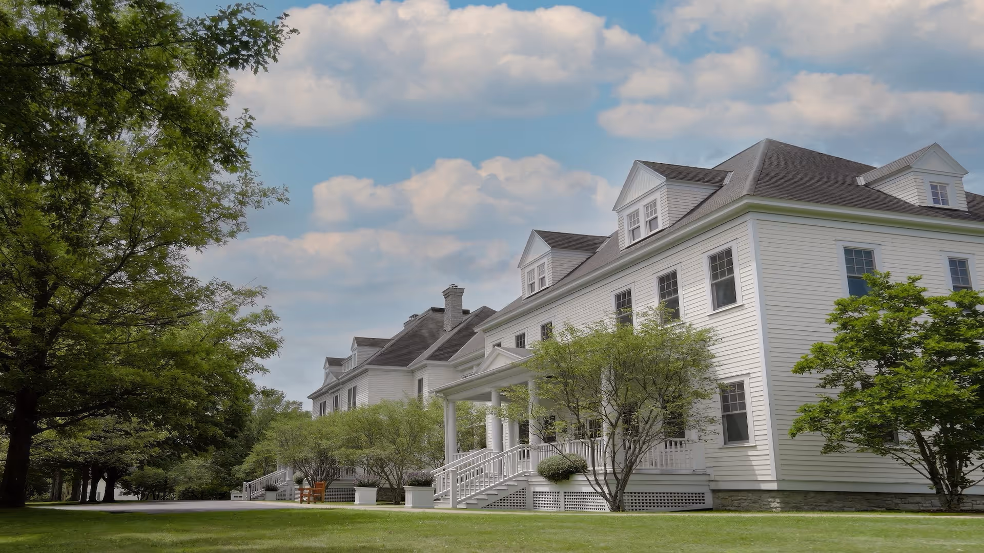 A large white multi-story building with a dark roof, surrounded by green trees and a well-maintained lawn under a partly cloudy sky.