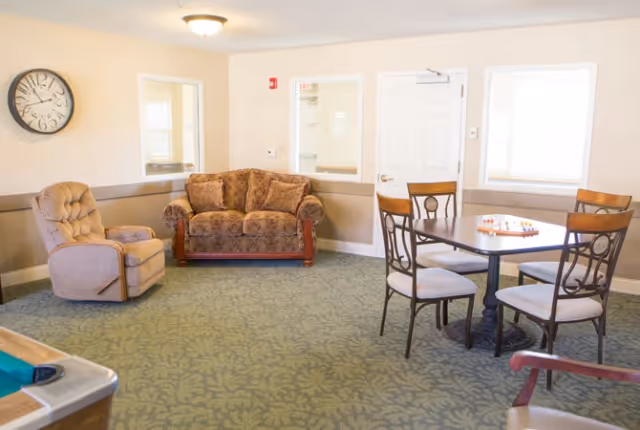A senior living facility common room with a patterned green carpet, a beige recliner chair, a brown upholstered loveseat, a square table with four chairs, and a chessboard on the table. The walls are light beige with two windows and a white door. A round wall clock is mounted on the left wall.