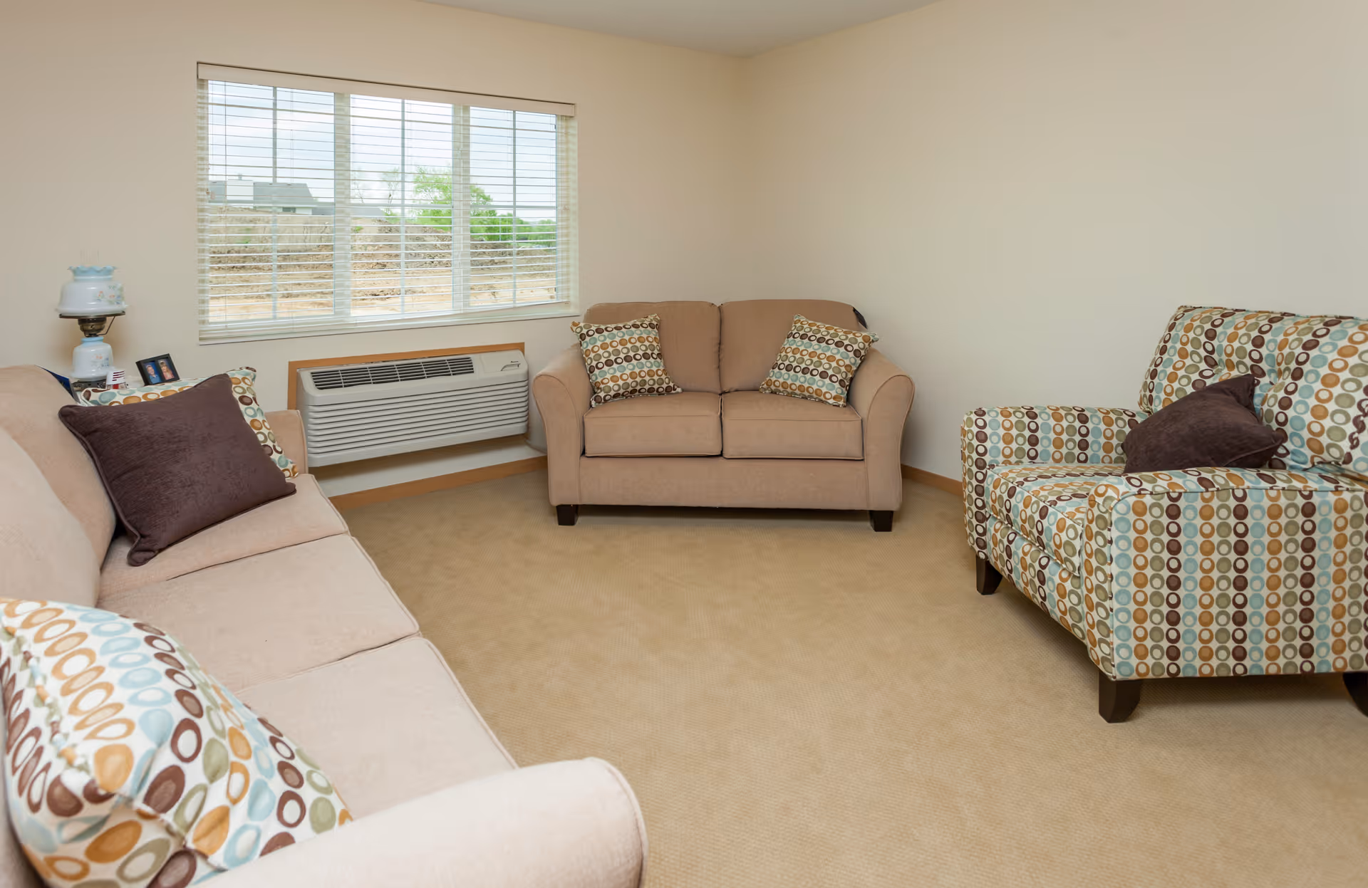 A cozy living room with beige carpet and light beige walls. The room contains a beige loveseat with two patterned pillows, a beige sofa with patterned and solid brown pillows, and a patterned armchair with a solid brown pillow. A window with white blinds is above a wall-mounted air conditioning unit. A small table with a vintage lamp and framed photos is next to the sofa.