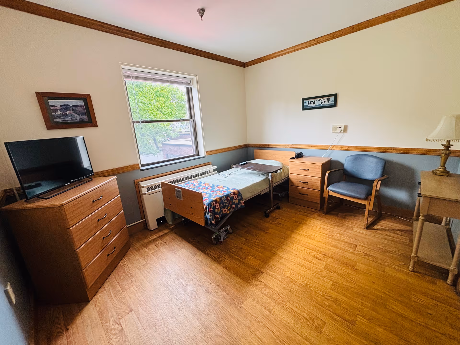 A small, simple bedroom in a rehabilitation and nursing center featuring a single hospital-style bed with a colorful blanket, a wooden dresser with a flat-screen TV on top, a blue upholstered chair, a wooden nightstand with a telephone, a small table lamp on a side table, and a window showing green trees outside. The room has wooden flooring and light-colored walls with wooden trim.