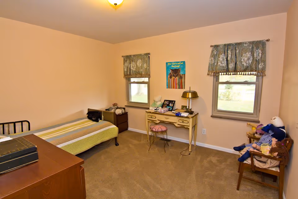 A small bedroom with beige walls and carpet, featuring a single bed with a striped bedspread, a wooden dresser, a vintage-style desk with a lamp and chair, two windows with floral curtains, and a wooden chair holding several dolls.