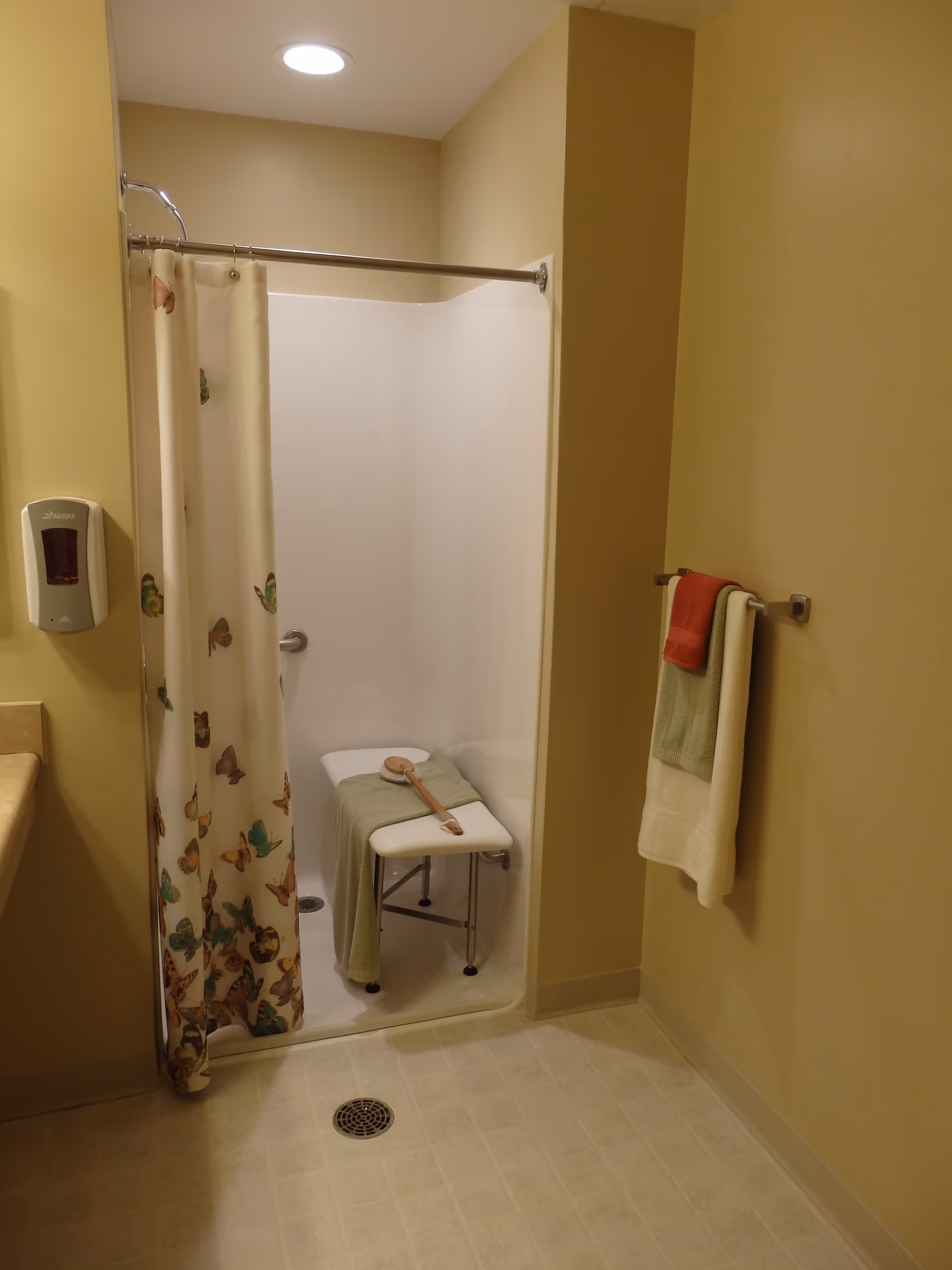 A bathroom with a walk-in shower featuring a butterfly-patterned shower curtain. Inside the shower is a white shower bench with a green towel and a long-handled bath brush on it. There is a towel rack on the wall outside the shower holding three towels in red, green, and white. A soap dispenser is mounted on the wall near the shower.
