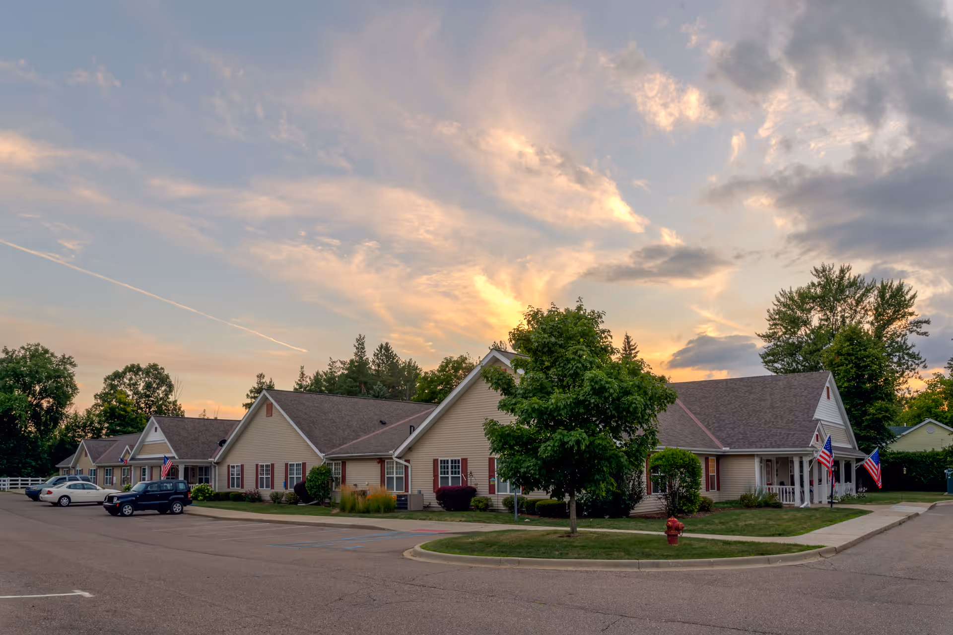 Exterior view of a single-story senior living facility building with beige siding and brown roofs, surrounded by trees and a parking lot with a few cars. The sky is partly cloudy with a warm sunset glow.