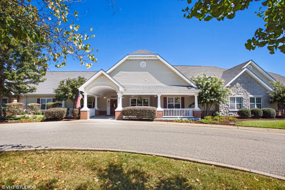Front exterior view of a single-story senior living facility building with a covered entrance, white siding, brick accents, and a stone section on the right. The building is surrounded by neatly trimmed bushes, trees, and a curved driveway under a clear blue sky.