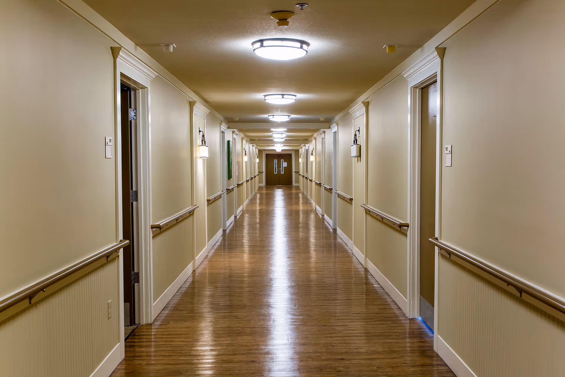 A long, well-lit hallway in an assisted living facility with wooden floors, beige walls, handrails on both sides, and multiple doors leading to rooms. Ceiling lights and wall sconces provide illumination.