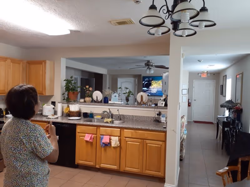 A woman stands in a kitchen area of an assisted living facility, facing away from the camera. The kitchen has wooden cabinets, a granite countertop with a sink, and various kitchen items including a rice cooker, bananas, and dish towels. Beyond the kitchen is a living room area with plants, decorative plates, and a television mounted on the wall. A hallway with a white door and exit sign is visible in the background.