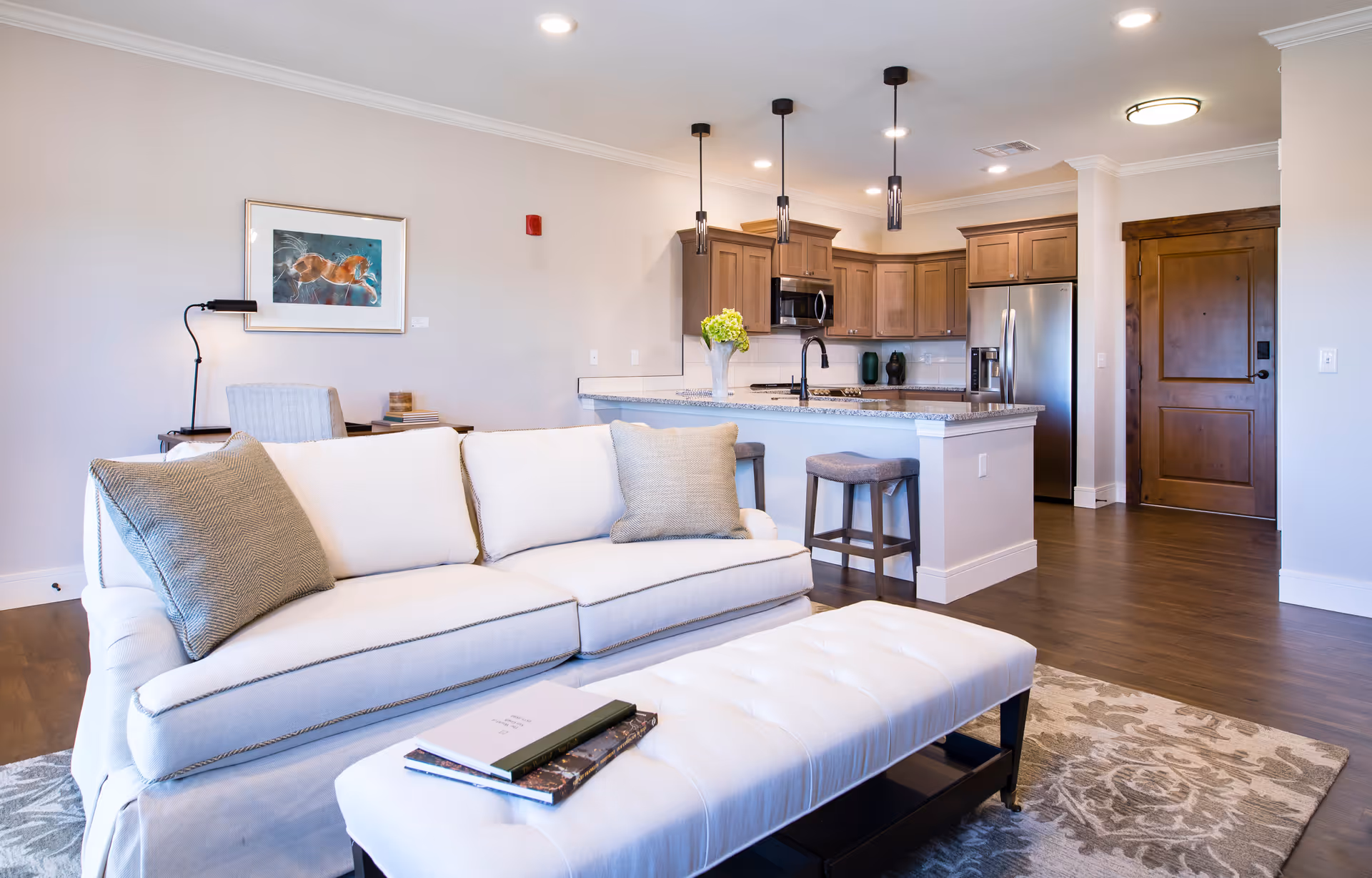 A bright and modern living room with a white sofa adorned with two gray pillows, a matching white ottoman with books on top, and a patterned area rug. In the background, there is an open kitchen with wooden cabinets, a granite countertop island with two stools, stainless steel appliances, and three pendant lights hanging from the ceiling. A wooden door and a framed picture on the wall are also visible.