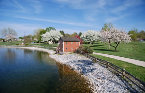 A peaceful outdoor scene at Menno Haven-Penn Hall featuring a small brown building near the edge of a calm lake with clear water. The shoreline is lined with white rocks and a wooden fence. In the background, there is a green grassy area with several blossoming trees under a blue sky with light clouds.