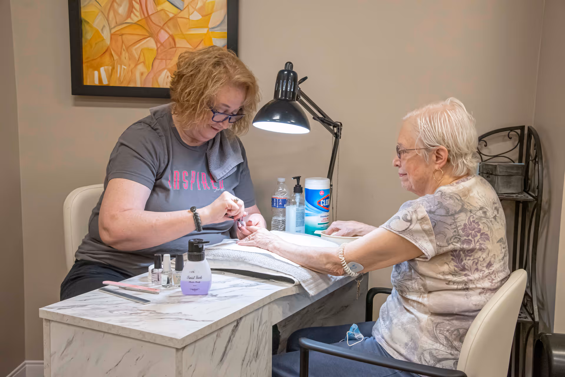 A woman is giving a manicure to an elderly woman seated at a small marble-patterned table. Various manicure supplies, including nail polish and hand lotion, are on the table. A lamp illuminates the workspace, and a colorful abstract painting hangs on the wall behind them.