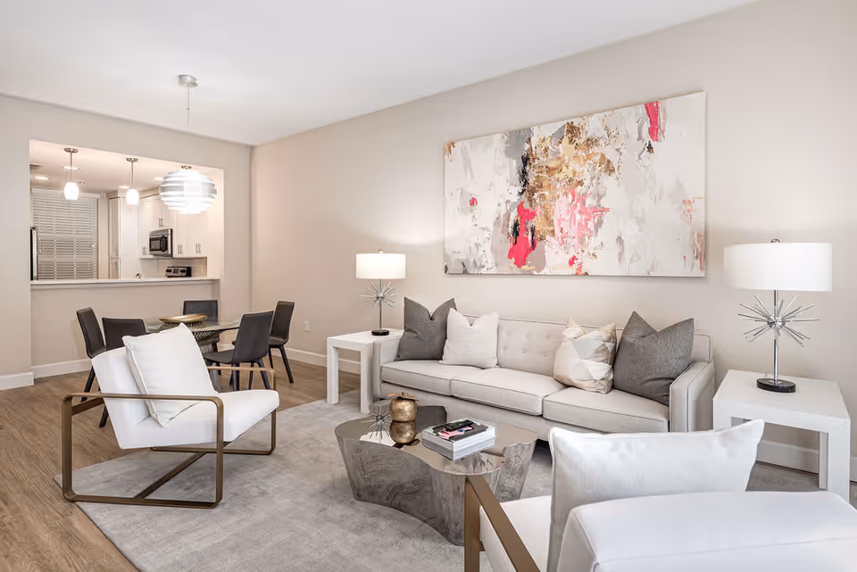 A modern living room with a light gray sofa adorned with various pillows, two white armchairs with metal frames, a reflective coffee table with books and decorative items, two white side tables each with a lamp, and a large abstract painting with pink, beige, and gray tones on the wall. In the background, there is a dining area with a glass table and four dark chairs, and a kitchen with white cabinetry and stainless steel appliances.