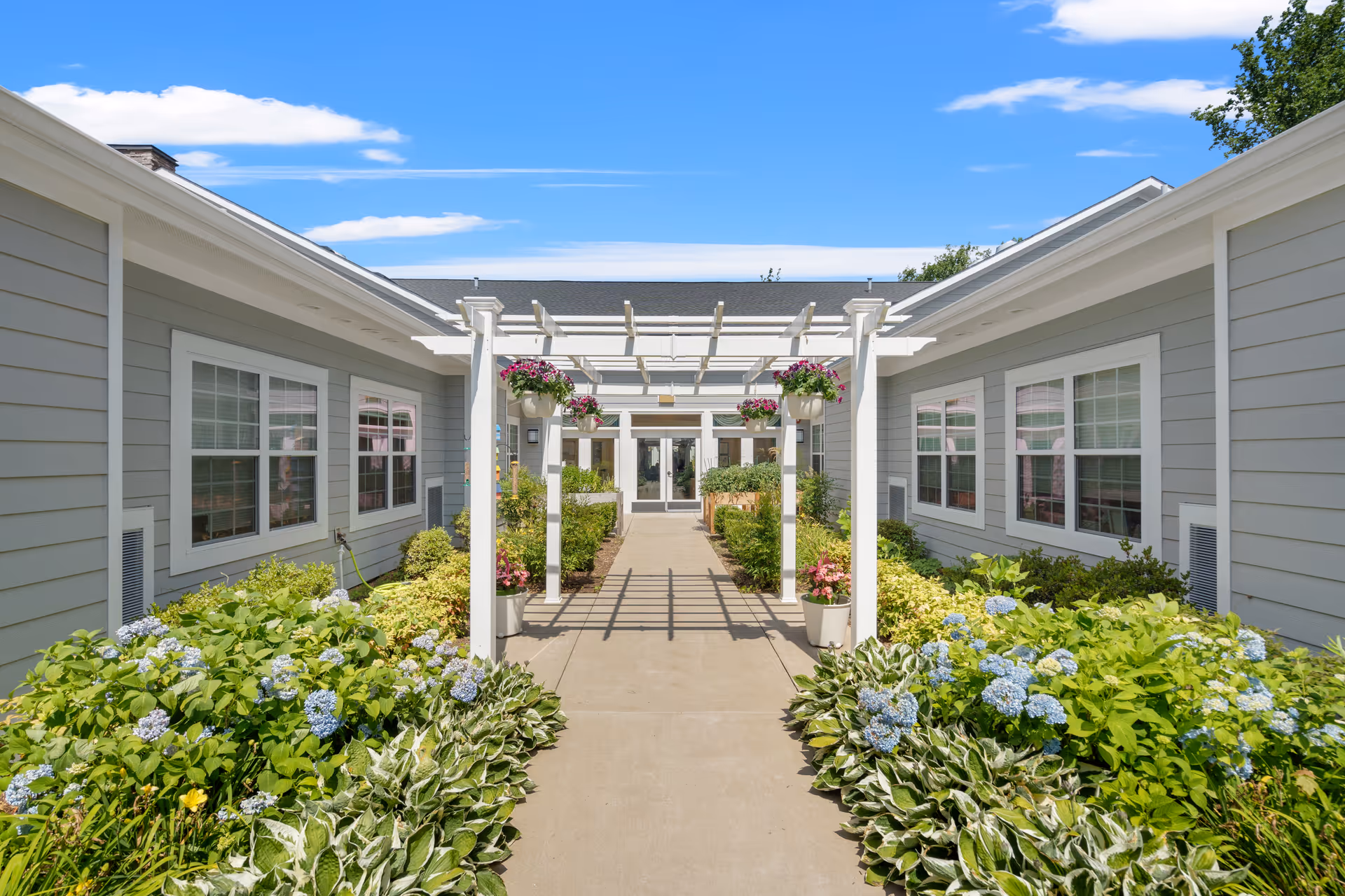 Outdoor walkway with a white pergola adorned with hanging flower pots leading to the entrance of a building. The walkway is flanked by lush green plants and blooming flowers, with light gray siding and white-trimmed windows on both sides of the building under a bright blue sky with scattered clouds.