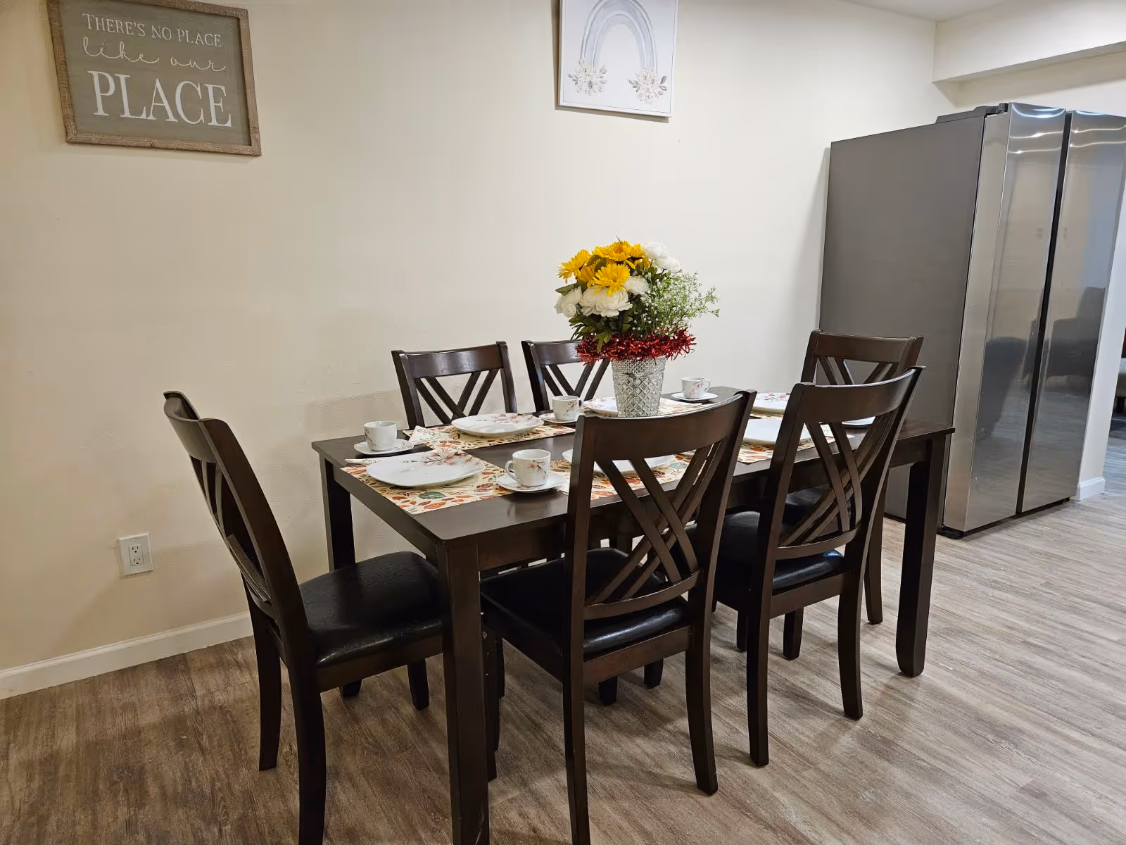 Wood dining table set for six with place settings and a floral centerpiece in a dining area next to a stainless steel refrigerator.