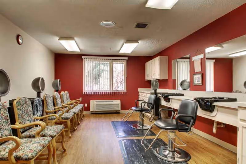 Interior salon room with styling chairs, hairdryers, mirrors and a row of floral-upholstered waiting chairs.