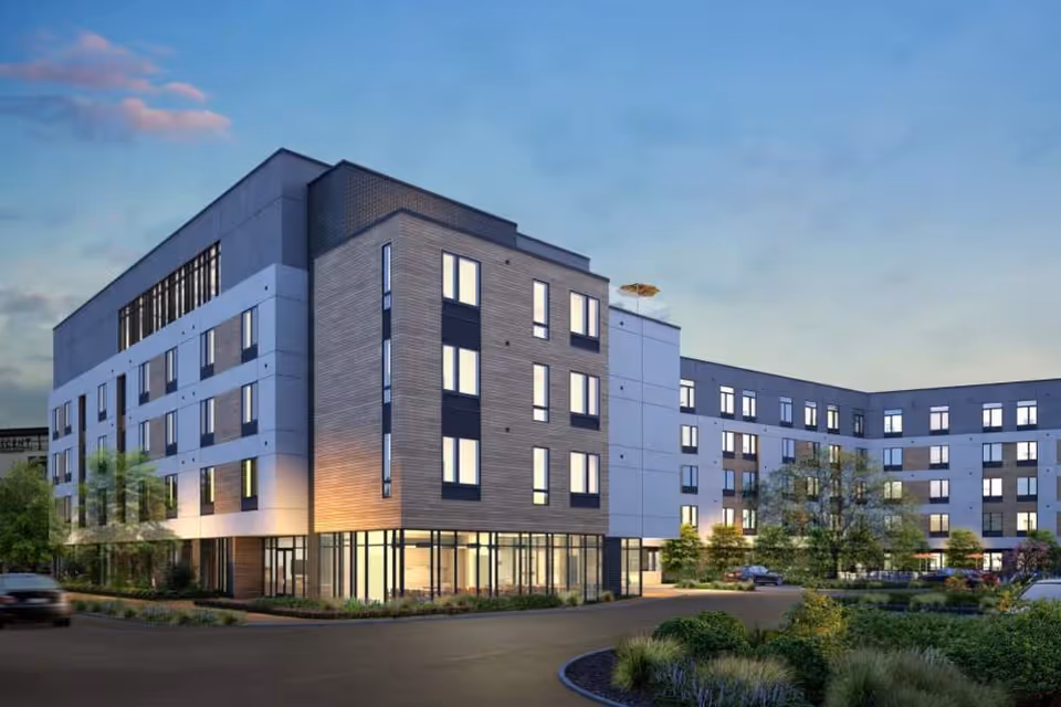 Exterior view of a modern four-story senior living building at dusk with lit windows, landscaping, and a driveway.