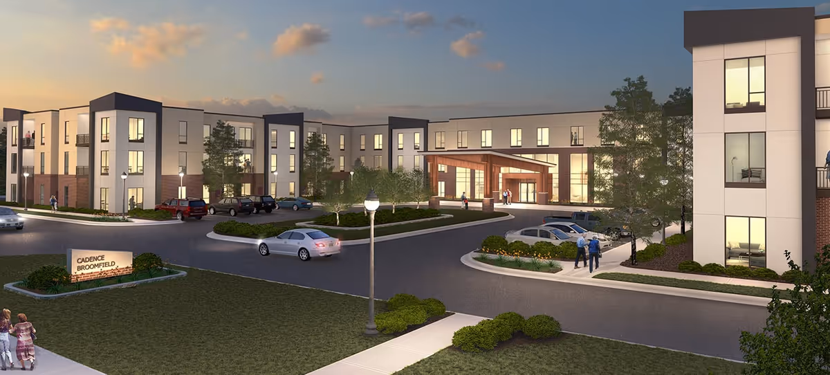 Exterior view of a modern three-story senior living facility at dusk with cars parked in front and people walking on the sidewalk. The building has large windows and a covered entrance with warm lighting. There is a sign on the lawn that reads 'Cadence Broomfield'.