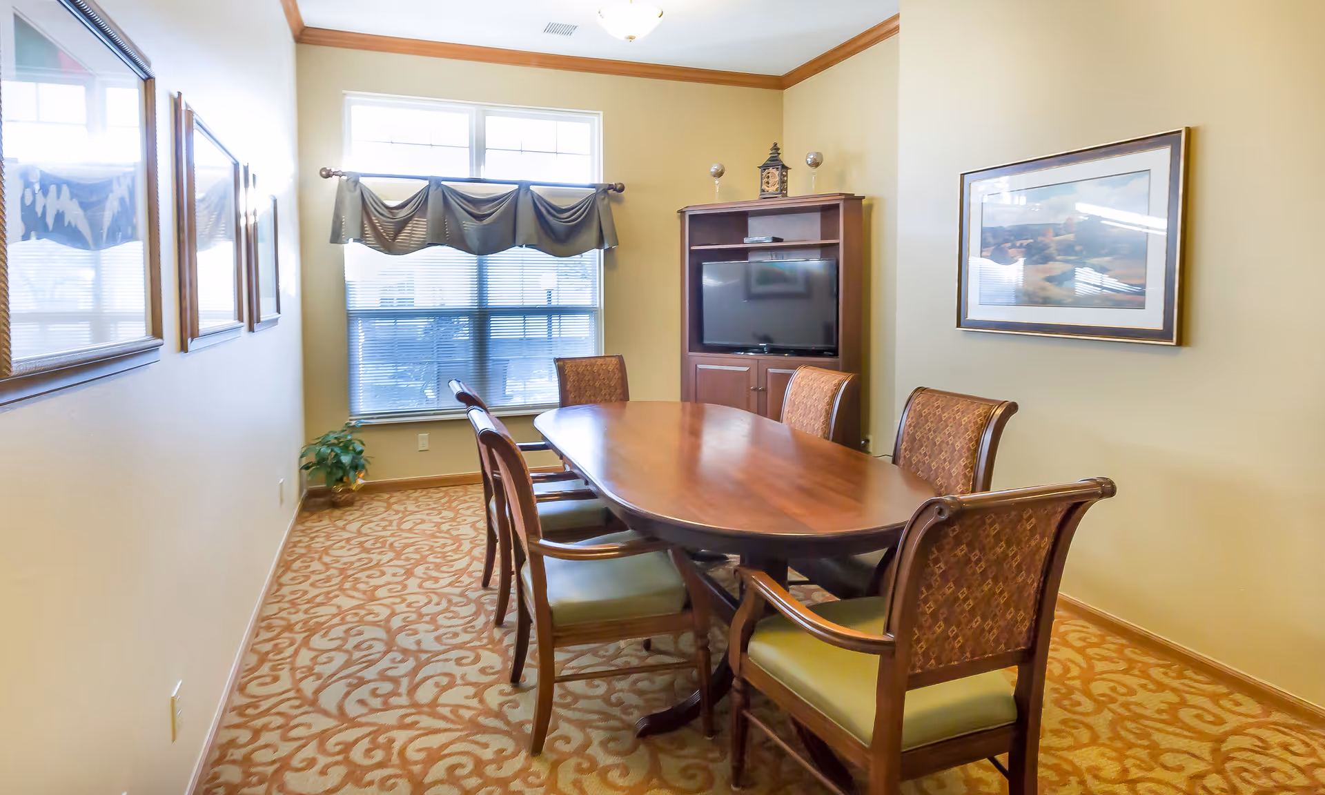 A dining room with a wooden oval table surrounded by six chairs with patterned upholstery and green cushions. The room has beige walls, a patterned carpet, a window with blinds and a valance, a wooden cabinet with a flat-screen TV, and framed artwork on the walls.