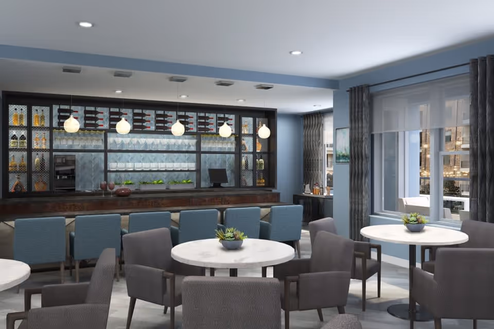 A modern dining area with round white tables and gray upholstered chairs. In the background, there is a bar with blue cushioned bar stools, shelves stocked with bottles and glasses, and pendant lights hanging from the ceiling. Large windows with gray curtains allow natural light to enter the room.