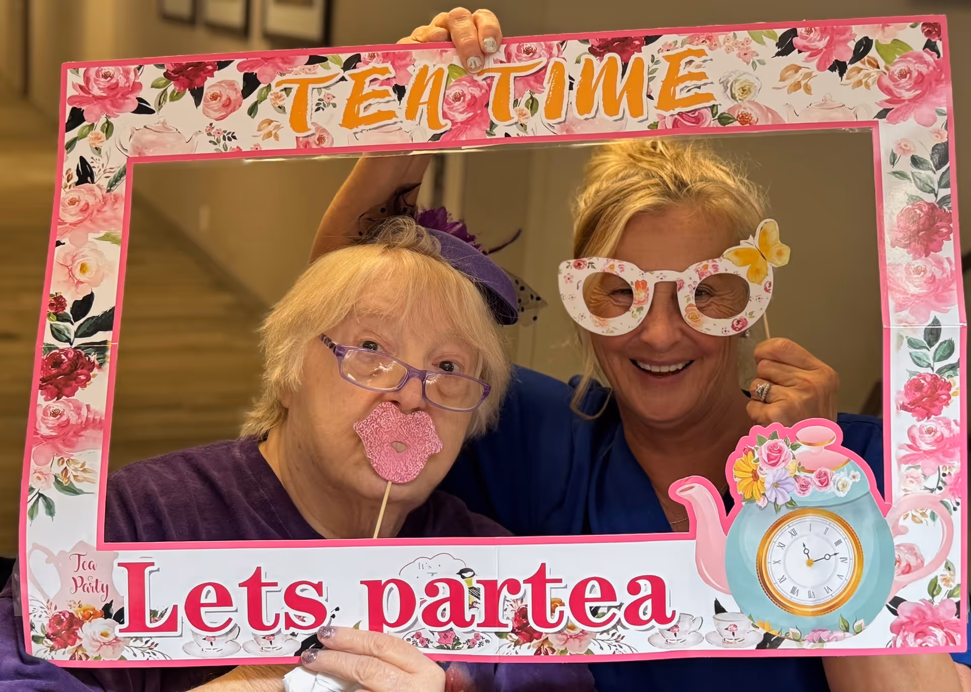 Two women posing behind a decorative photo frame that says 'TEN TIME Lets partea' with floral designs and a teapot illustration. One woman is wearing purple glasses and holding a pink lips prop, while the other is wearing floral paper glasses and smiling.